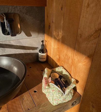 Foot with red nail polish in a floral bag on a wooden surface next to a bottle of soap. Buttermilk Grace Carry All Bag Secret Garden - The Rose Edit