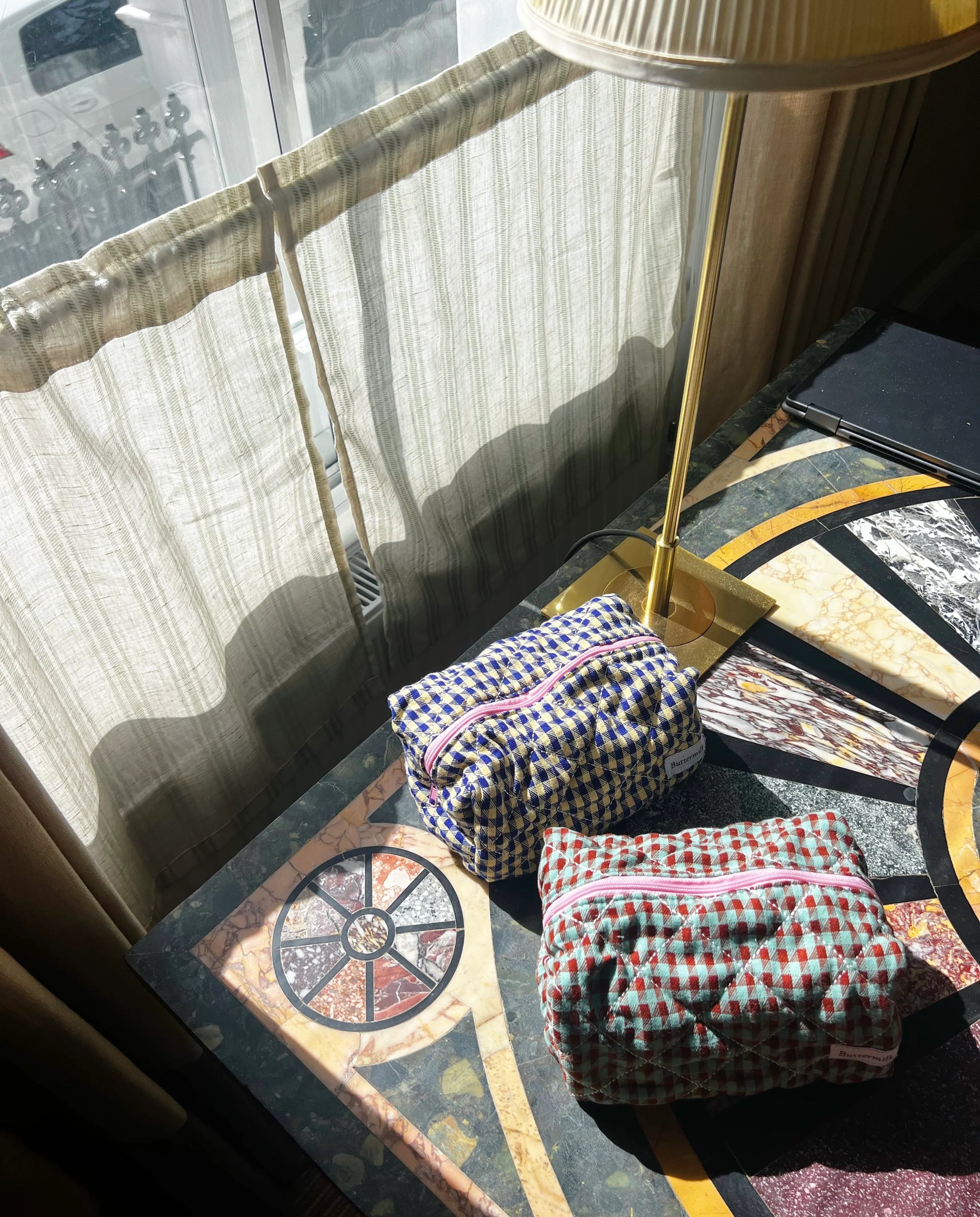 Two checkered bags on a decorative table with a lamp and curtains in the background. Buttermilk Grace Carry All Bag Boysenberry Pie - The Rose Edit
