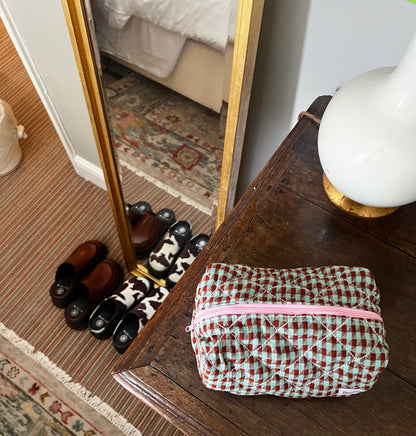 Quilted toiletry bag with pink zipper on a wooden surface, with shoes and a mirror in the background. Buttermilk Grace Carry All Bag Boysenberry Pie - The Rose Edit