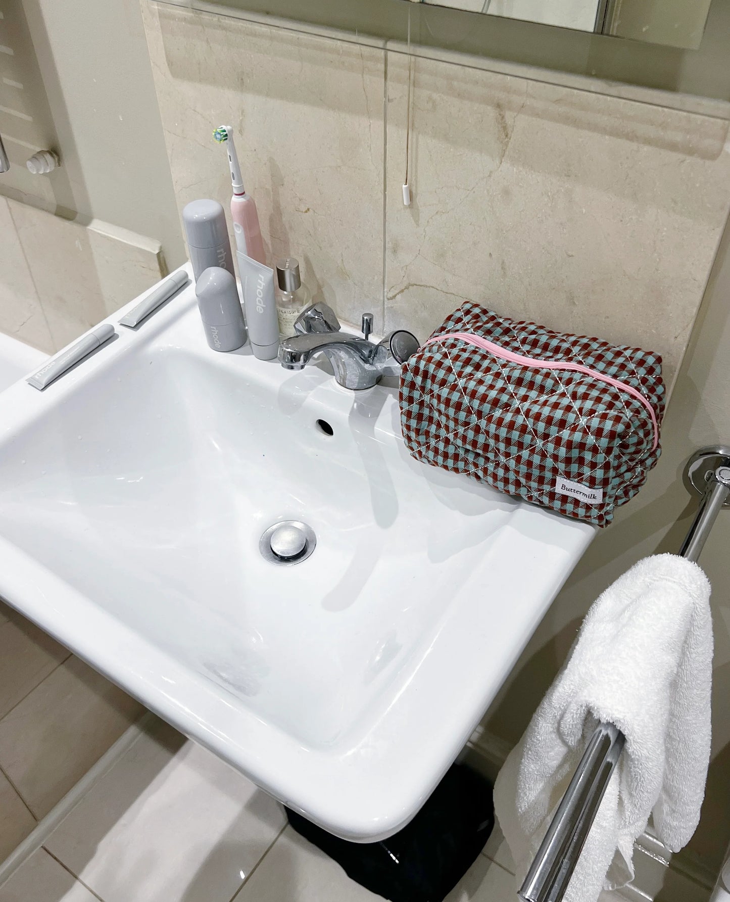 Bathroom sink with toiletries and a checkered bag on a towel. Buttermilk Grace Carry All Bag Boysenberry Pie - The Rose Edit