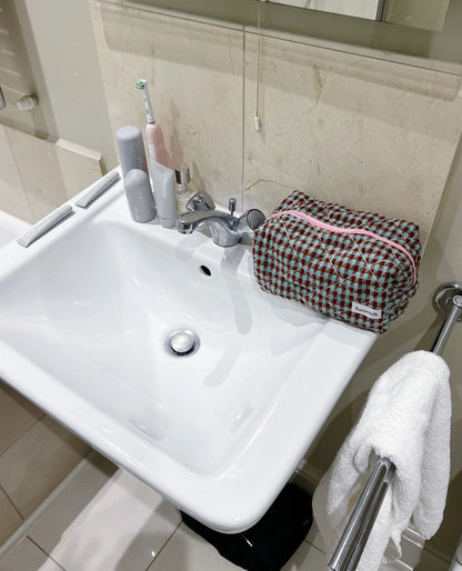 Bathroom sink with toiletries and a checkered bag on a towel. Buttermilk Grace Carry All Bag Boysenberry Pie - The Rose Edit