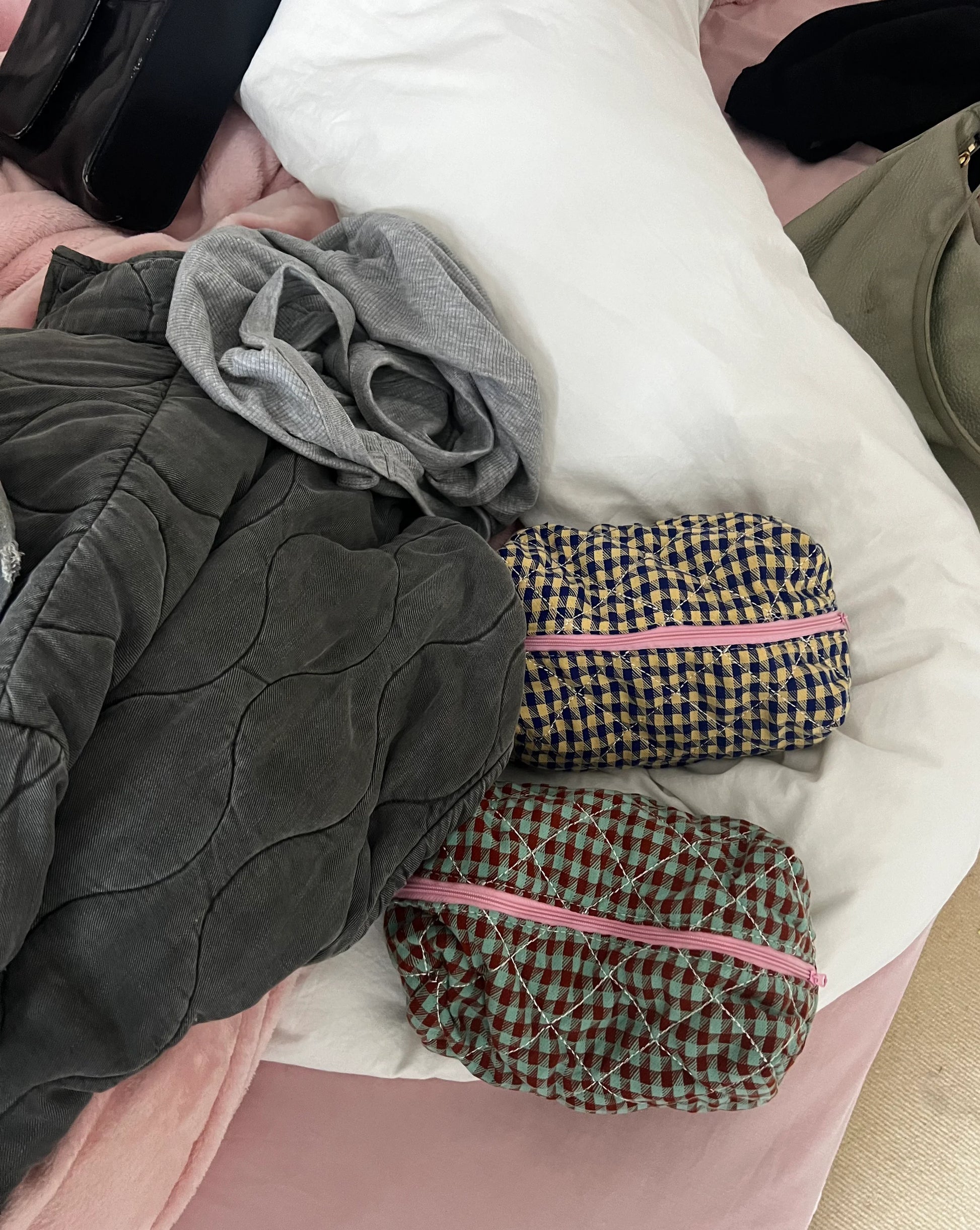 Two checkered pouches with pink zippers on a bed with gray and white bedding. Buttermilk Grace Carry All Bag Boysenberry Pie - The Rose Edit