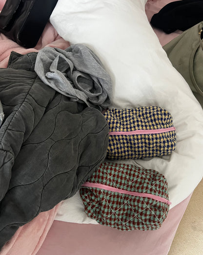 Two checkered pouches with pink zippers on a bed with gray and white bedding. Buttermilk Grace Carry All Bag Boysenberry Pie - The Rose Edit