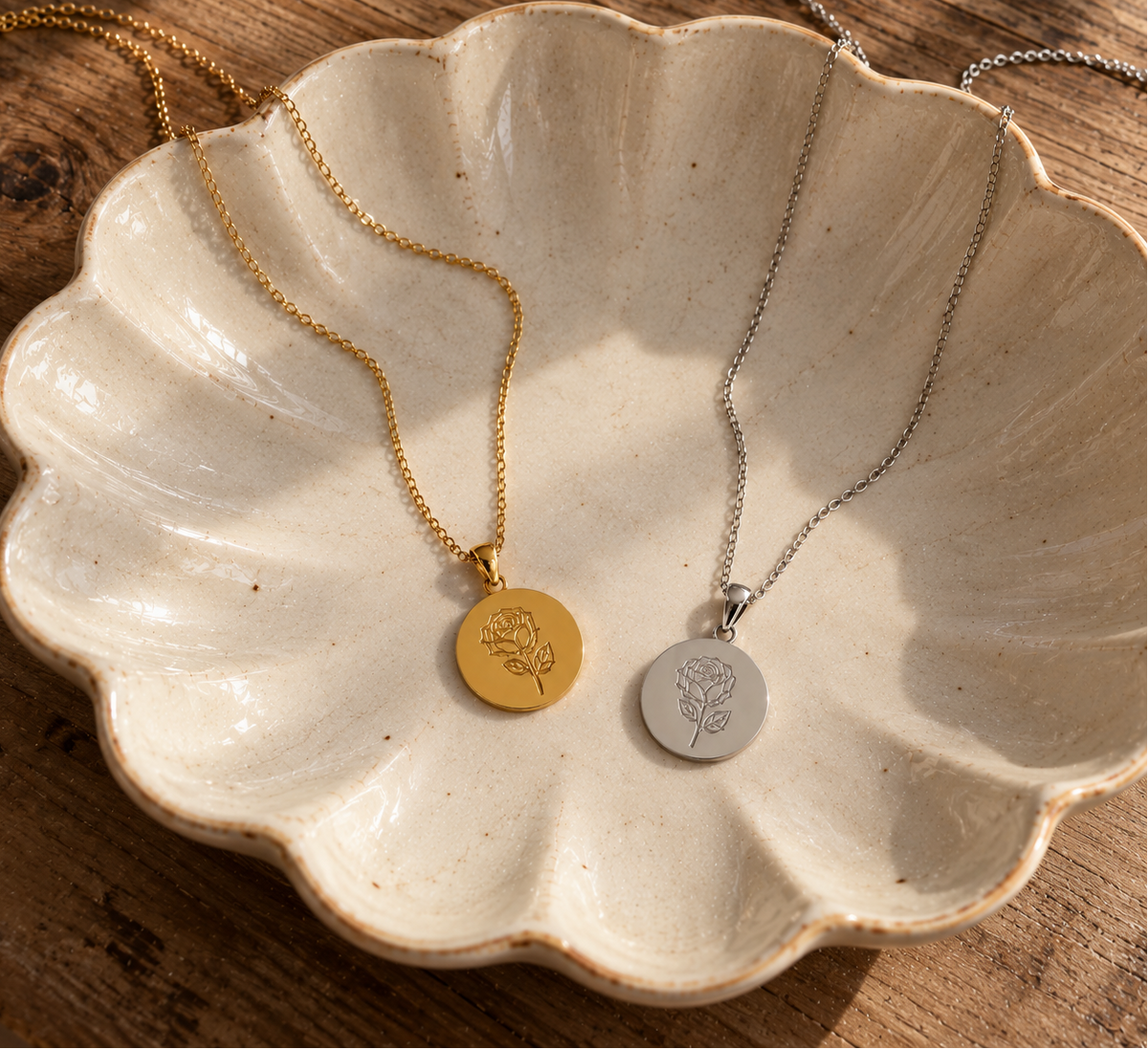 Two round pendant necklaces displayed in a scalloped ceramic dish on a wooden surface. One necklace is gold with a gold pendant engraved with a floral motif, and the other is silver with a silver pendant engraved with the same floral motif.