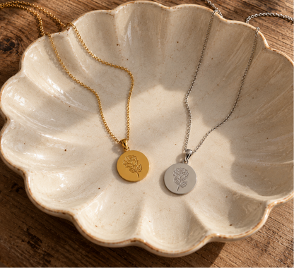 Two round pendant necklaces displayed in a scalloped ceramic dish on a wooden surface. One necklace is gold with a gold pendant engraved with a floral motif, and the other is silver with a silver pendant engraved with the same floral motif.