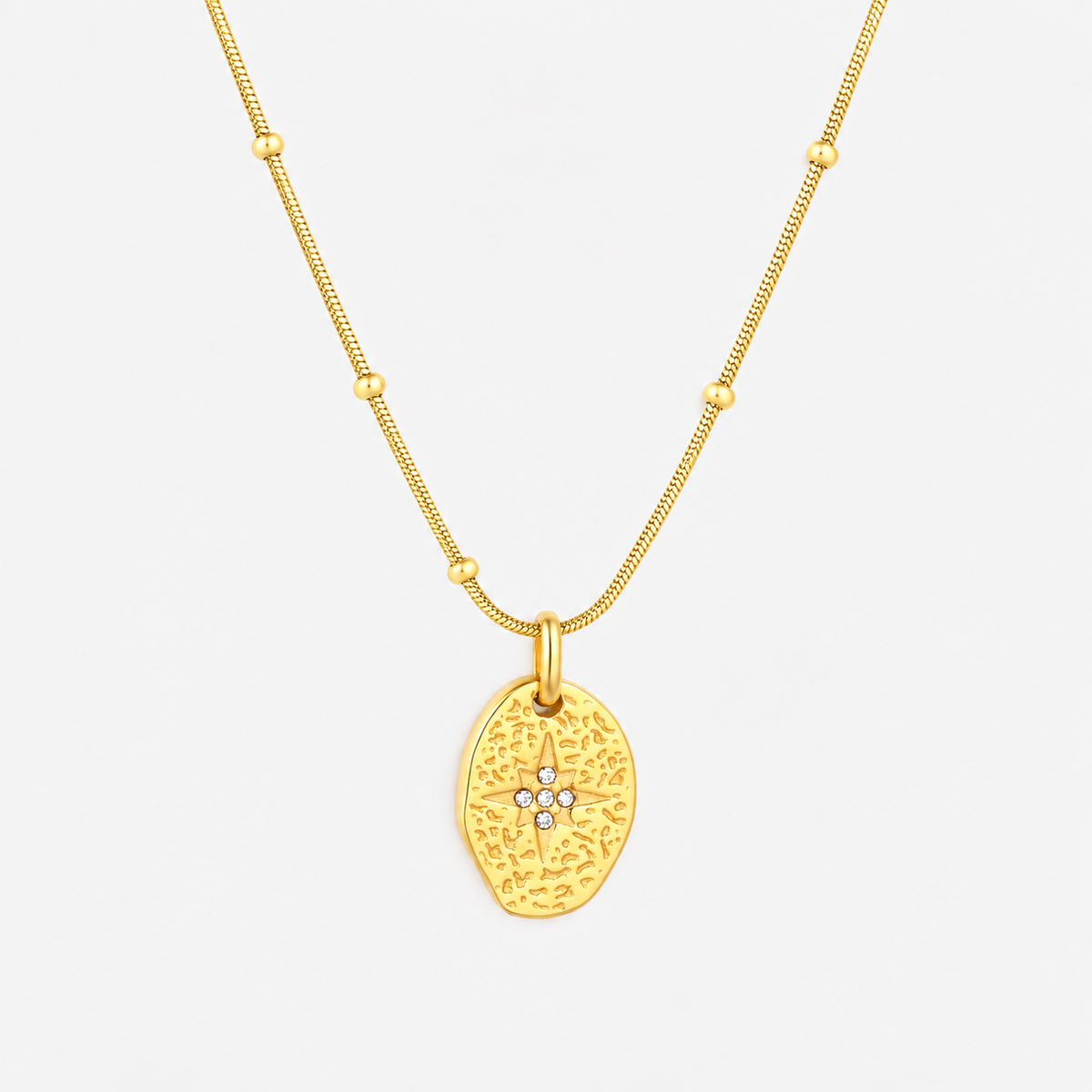 A gold necklace featuring a textured, organic-shaped pendant with a small star motif set with tiny clear stones at the center, suspended from a delicate beaded chain against a light gray background.