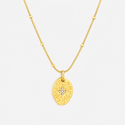 A gold necklace featuring a textured, organic-shaped pendant with a small star motif set with tiny clear stones at the center, suspended from a delicate beaded chain against a light gray background.