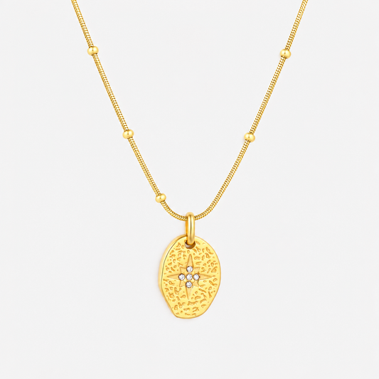 A gold necklace featuring a textured, organic-shaped pendant with a small star motif set with tiny clear stones at the center, suspended from a delicate beaded chain against a light gray background.