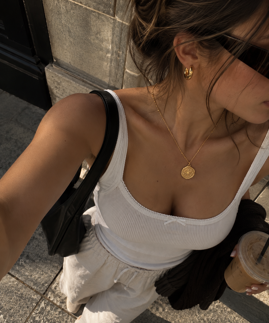 A woman wearing a delicate gold coin pendant necklace and matching gold hoop earring, styled with a white ribbed tank top and light-colored pants in an outdoor urban setting.
