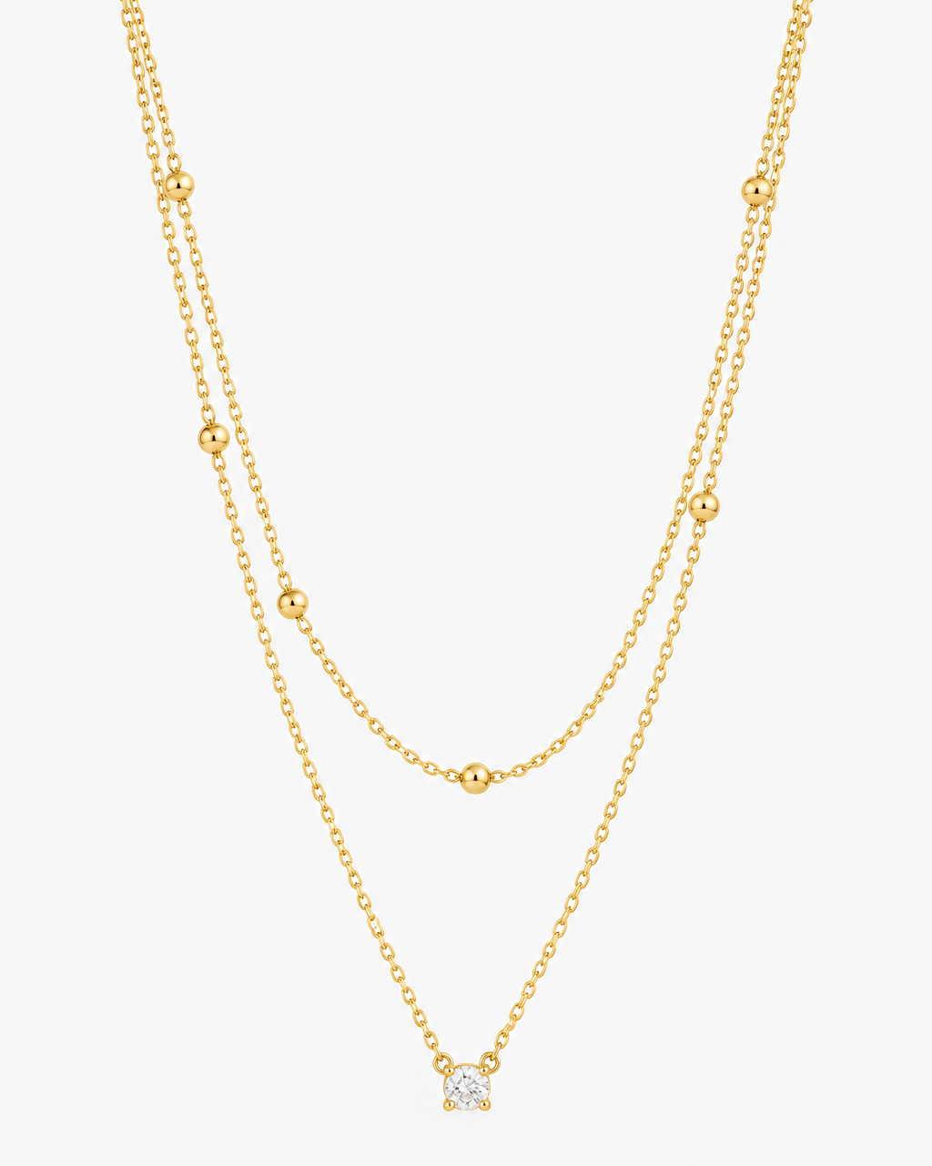 A delicate double-layered gold necklace featuring small bead accents on the upper layer and a small geometric diamond-shaped pendant on the lower layer, shown against a plain light gray background.