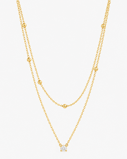 A delicate double-layered gold necklace featuring small bead accents on the upper layer and a small geometric diamond-shaped pendant on the lower layer, shown against a plain light gray background.