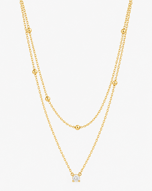 A delicate double-layered gold necklace featuring small bead accents on the upper layer and a small geometric diamond-shaped pendant on the lower layer, shown against a plain light gray background.