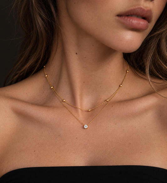 A close-up of a model wearing a delicate gold double-layer necklace with a small round clear gemstone pendant, styled with a black strap top against a dark background.