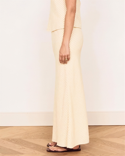 A model wearing a cream and beige vertically striped knit maxi skirt and matching top, paired with dark brown flat sandals, standing on a light wood herringbone floor against a white wall.