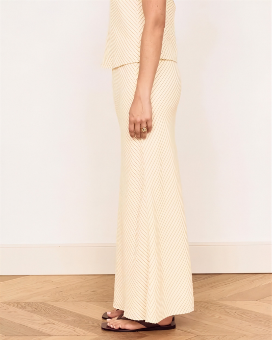 A model wearing a cream and beige vertically striped knit maxi skirt and matching top, paired with dark brown flat sandals, standing on a light wood herringbone floor against a white wall.