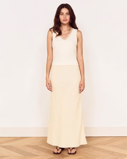 A woman modeling a sleeveless cream maxi dress with a textured, dotted pattern, standing against a plain off-white wall on a light wood herringbone floor.