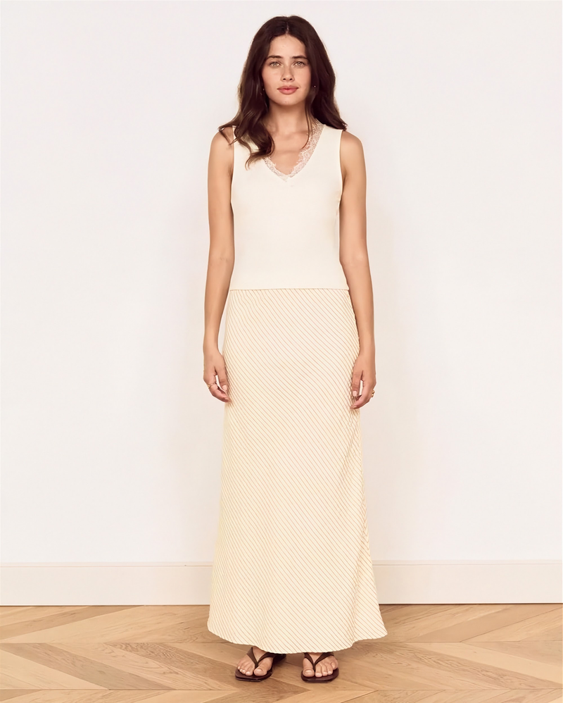 A woman modeling a sleeveless cream maxi dress with a textured, dotted pattern, standing against a plain off-white wall on a light wood herringbone floor.