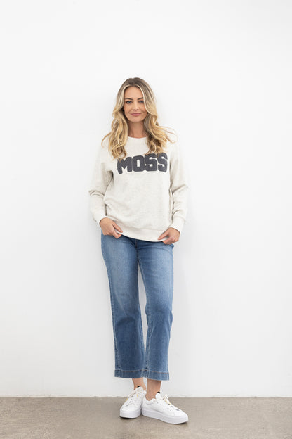 Woman wearing a cream sweatshirt with 'MOSS' text, blue jeans, and white sneakers on a plain background