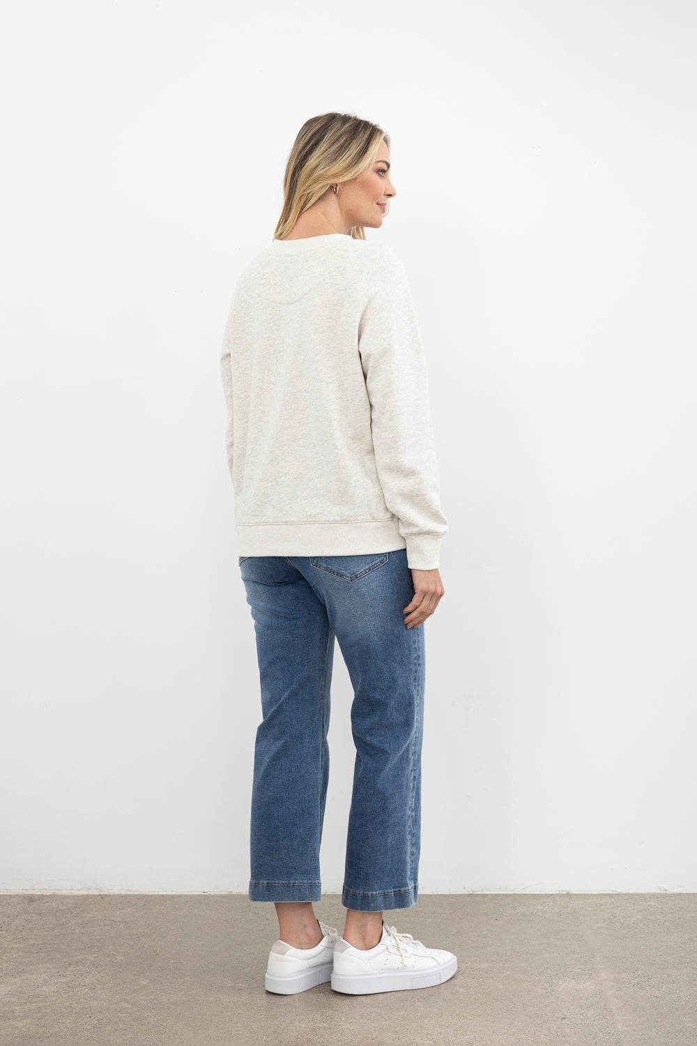 Person wearing a cream sweater and blue jeans standing against a white wall.