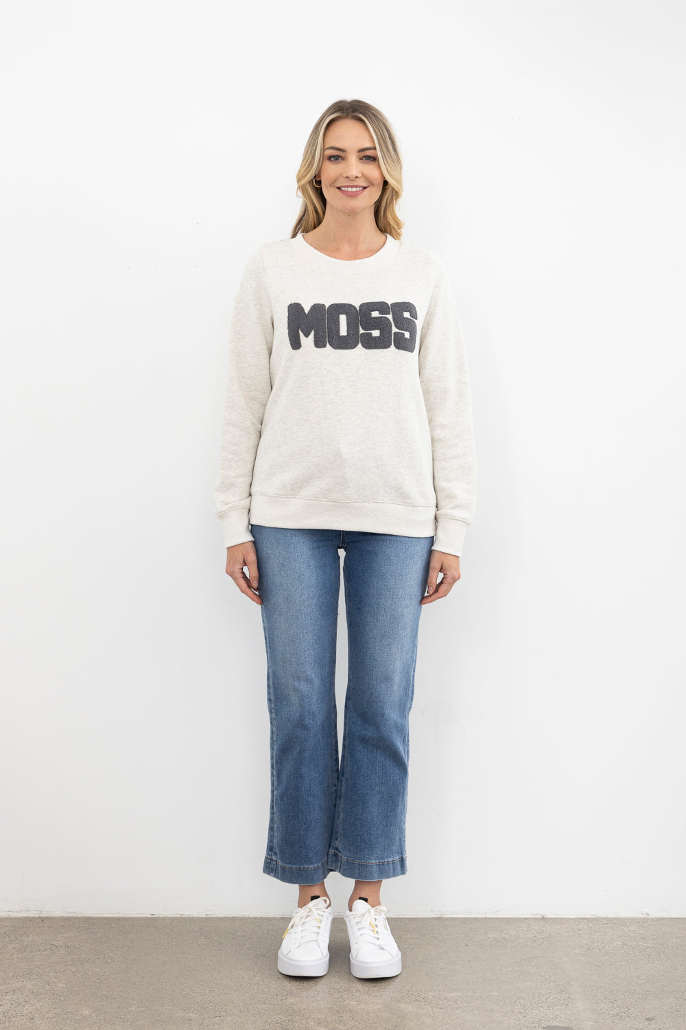 Woman wearing a white sweatshirt with 'MOSS' text and blue jeans on a white background