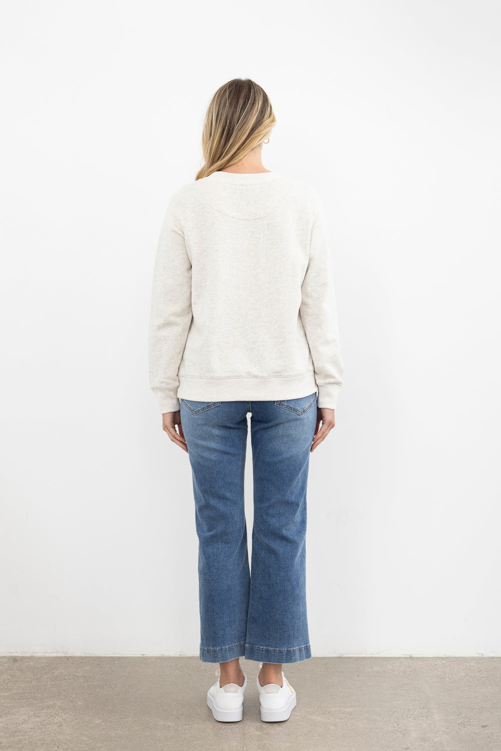 Person wearing a white sweater and blue jeans standing against a white wall.