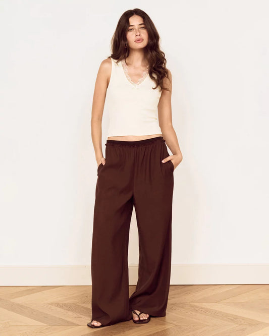 A woman modeling a sleeveless cream top and dark brown wide-leg pants, standing against a white wall on a light wood herringbone floor.