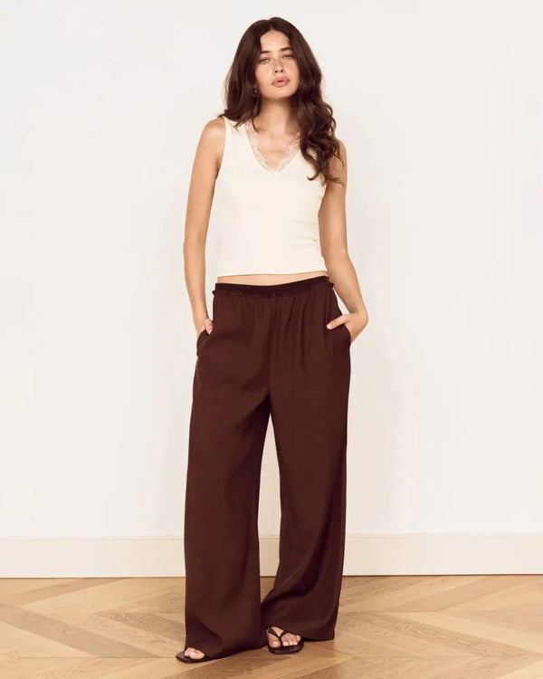 A woman modeling a sleeveless cream top and dark brown wide-leg pants, standing against a white wall on a light wood herringbone floor.