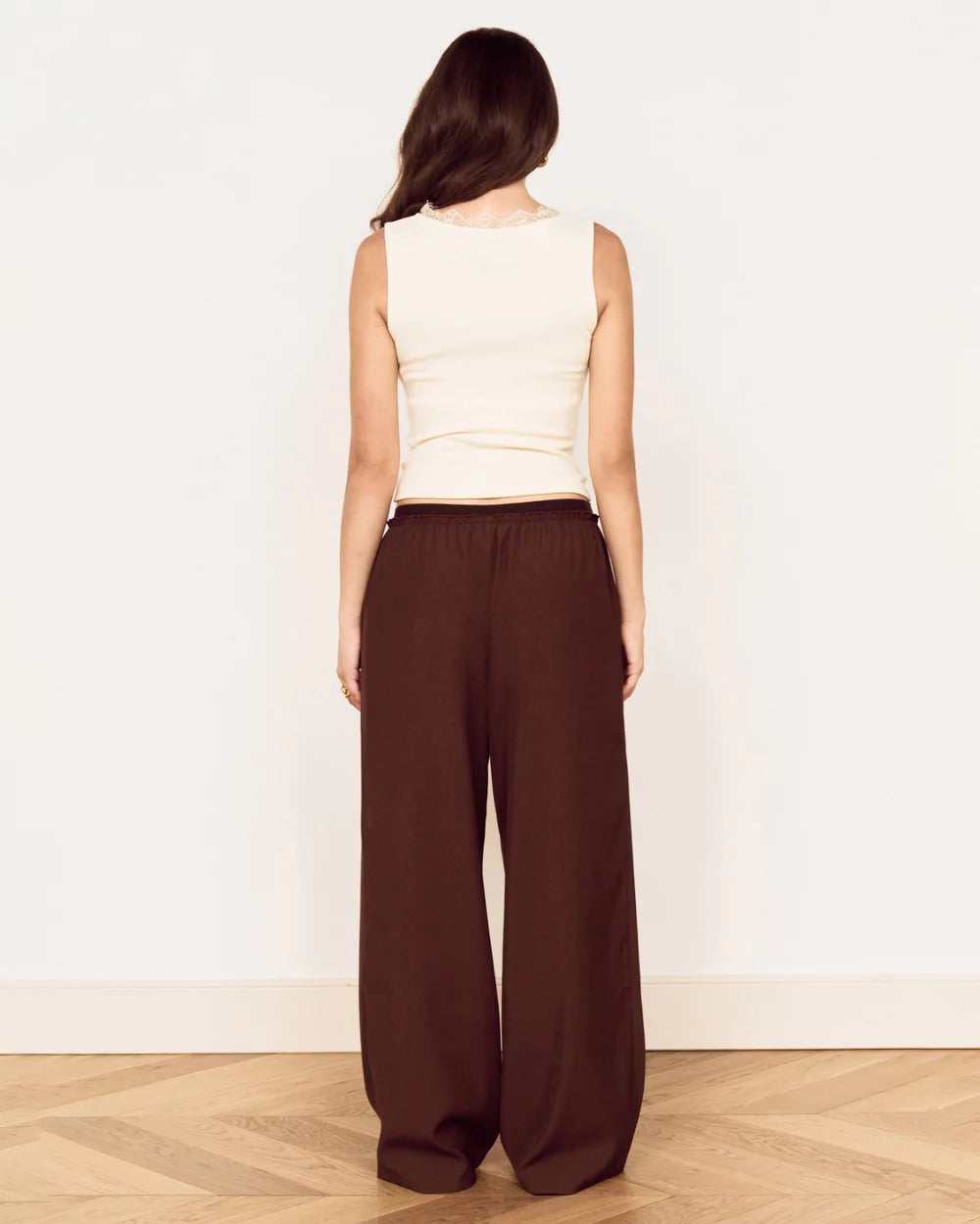 A woman shown from the back wearing a cream sleeveless top and dark brown wide-leg pants, standing on a light wood herringbone floor against a plain white wall.