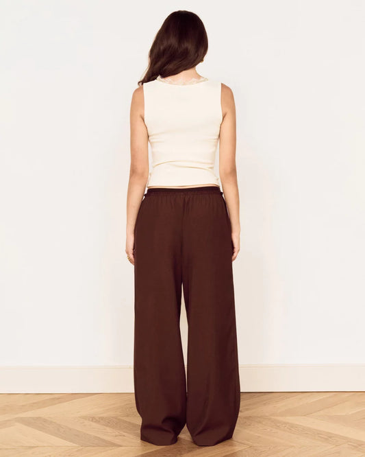 A woman shown from the back wearing a cream sleeveless top and dark brown wide-leg pants, standing on a light wood herringbone floor against a plain white wall.