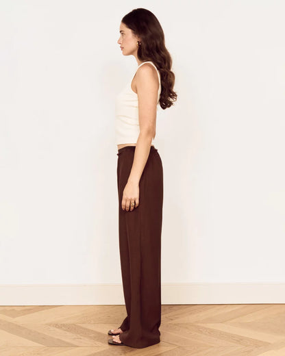 A woman in side profile wearing a white ribbed tank top and dark brown wide-leg trousers, standing on a light wood herringbone floor against a plain white wall.