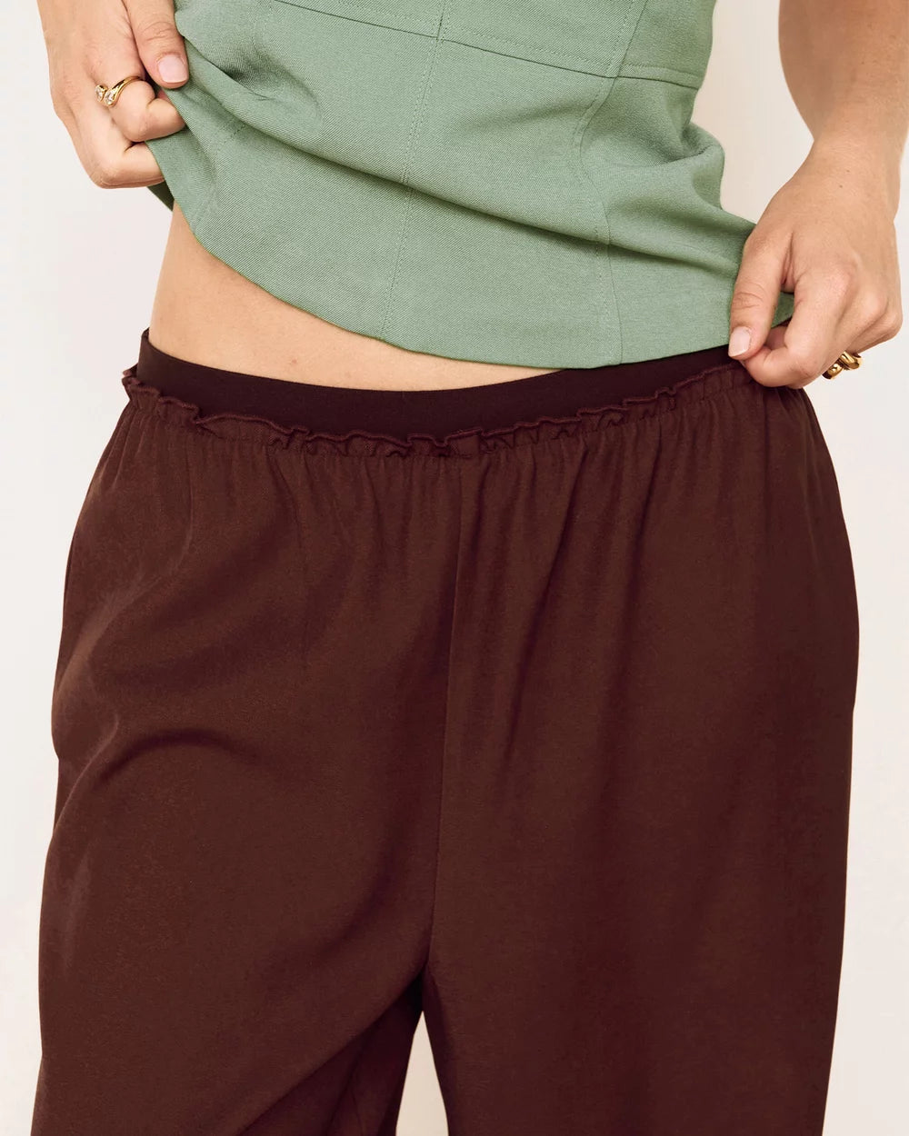 A close-up of a person wearing dark brown wide-leg pants with a ruffled elastic waistband, paired with a sage green top.
