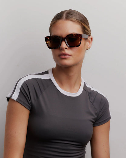 Woman wearing sunglasses and a gray athletic shirt with white accents against a plain background