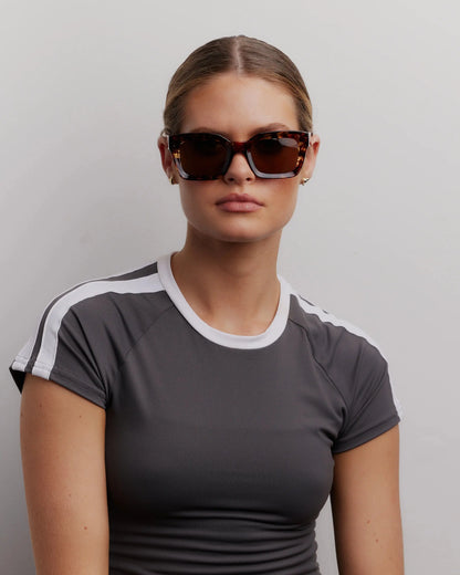 Woman wearing sunglasses and a gray athletic shirt with white accents against a plain background