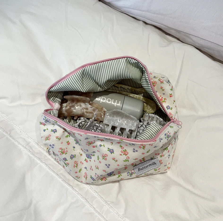 Floral-patterned bag with baby items on a white blanket. Buttermilk Grace Carry All Bag Forget-Me-Not - The Rose Edit