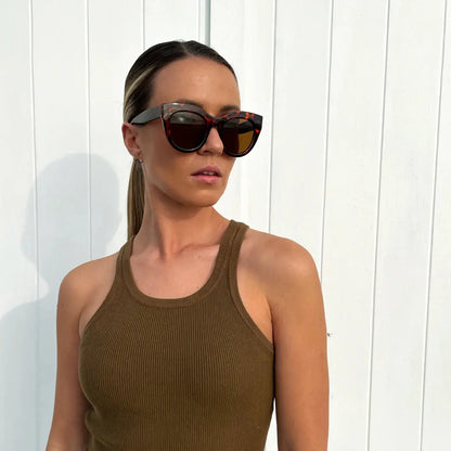 Woman wearing sunglasses and a brown tank top against a white wooden background