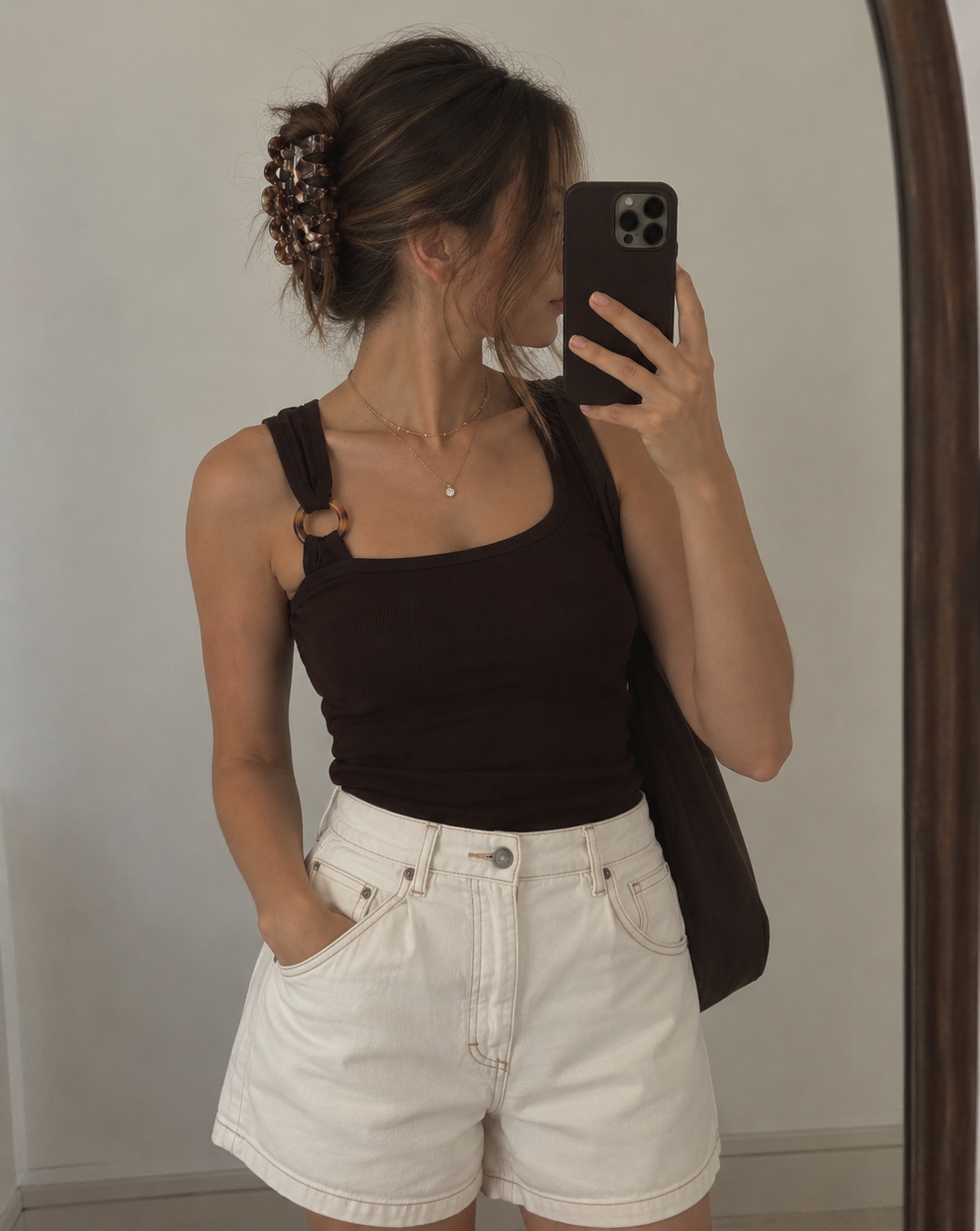 A woman taking a mirror selfie wearing a dark brown ribbed tank top with a unique ring detail on the strap, paired with white denim shorts.