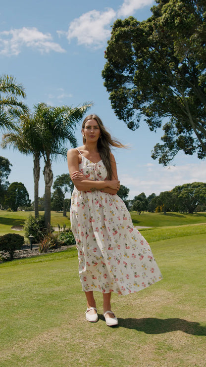 Woman in a floral dress standing on a grassy field with trees and blue sky in the background. Zinnia Dress Summer Haven Front View - The Rose Edit