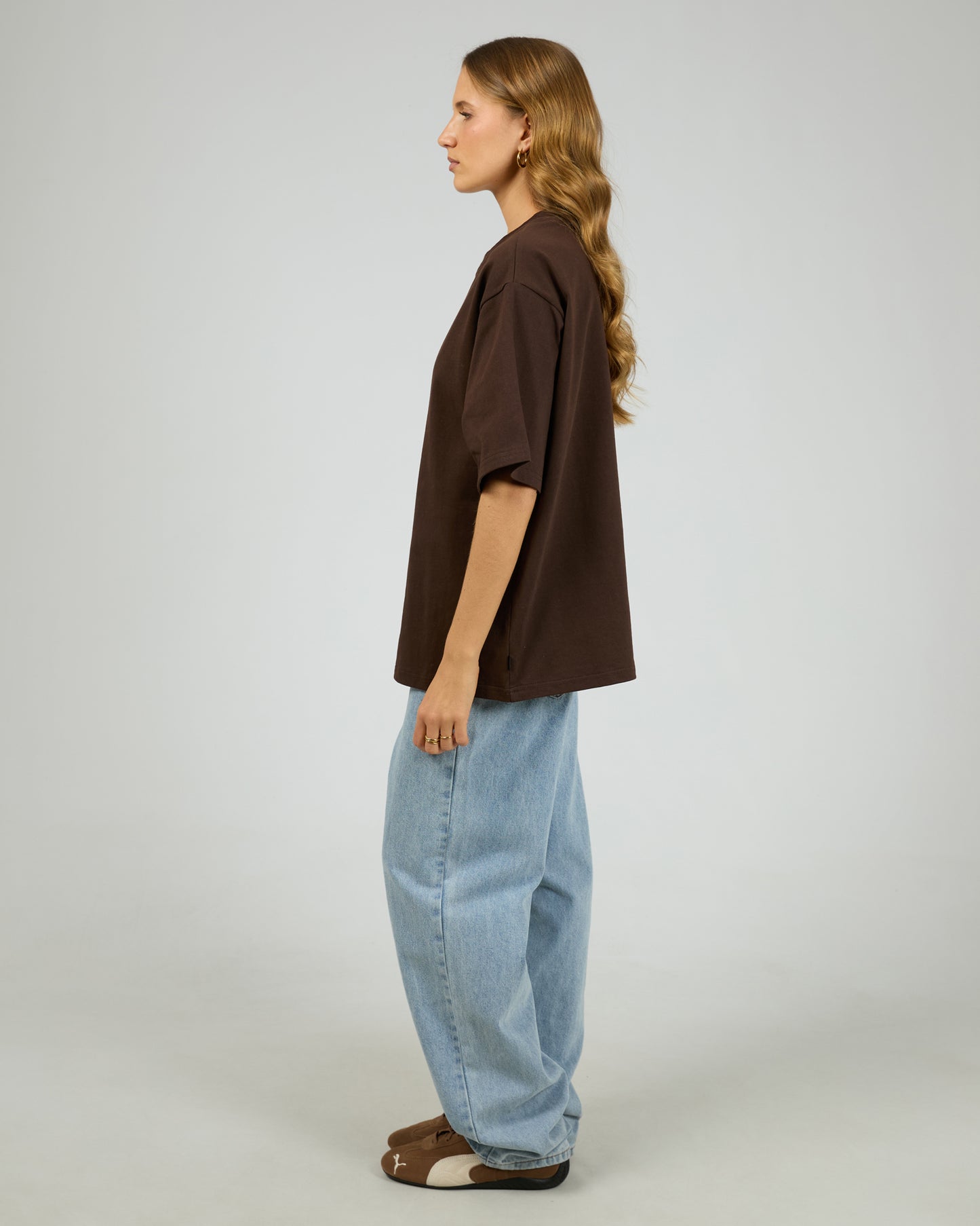 Person wearing a brown shirt and light blue jeans on a plain background. Core Oversized Tee Chocolate - The Rose Edit
