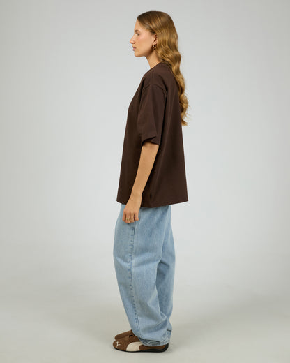 Person wearing a brown shirt and light blue jeans on a plain background. Core Oversized Tee Chocolate - The Rose Edit