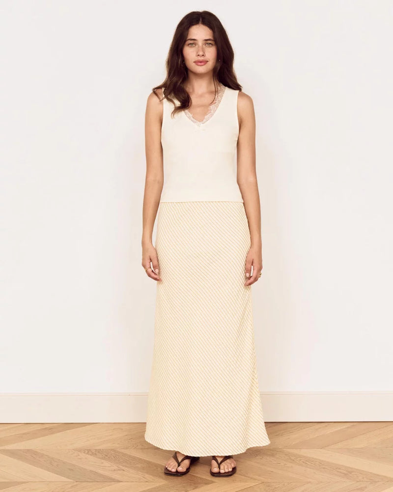 A woman modeling a sleeveless cream maxi dress with a textured pattern, standing against a plain white wall on a light wood herringbone floor.