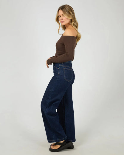 Woman wearing a brown top and blue jeans on a white background. Kylie Off Shoulder L/S Top Brown Side View - The Rose Edit